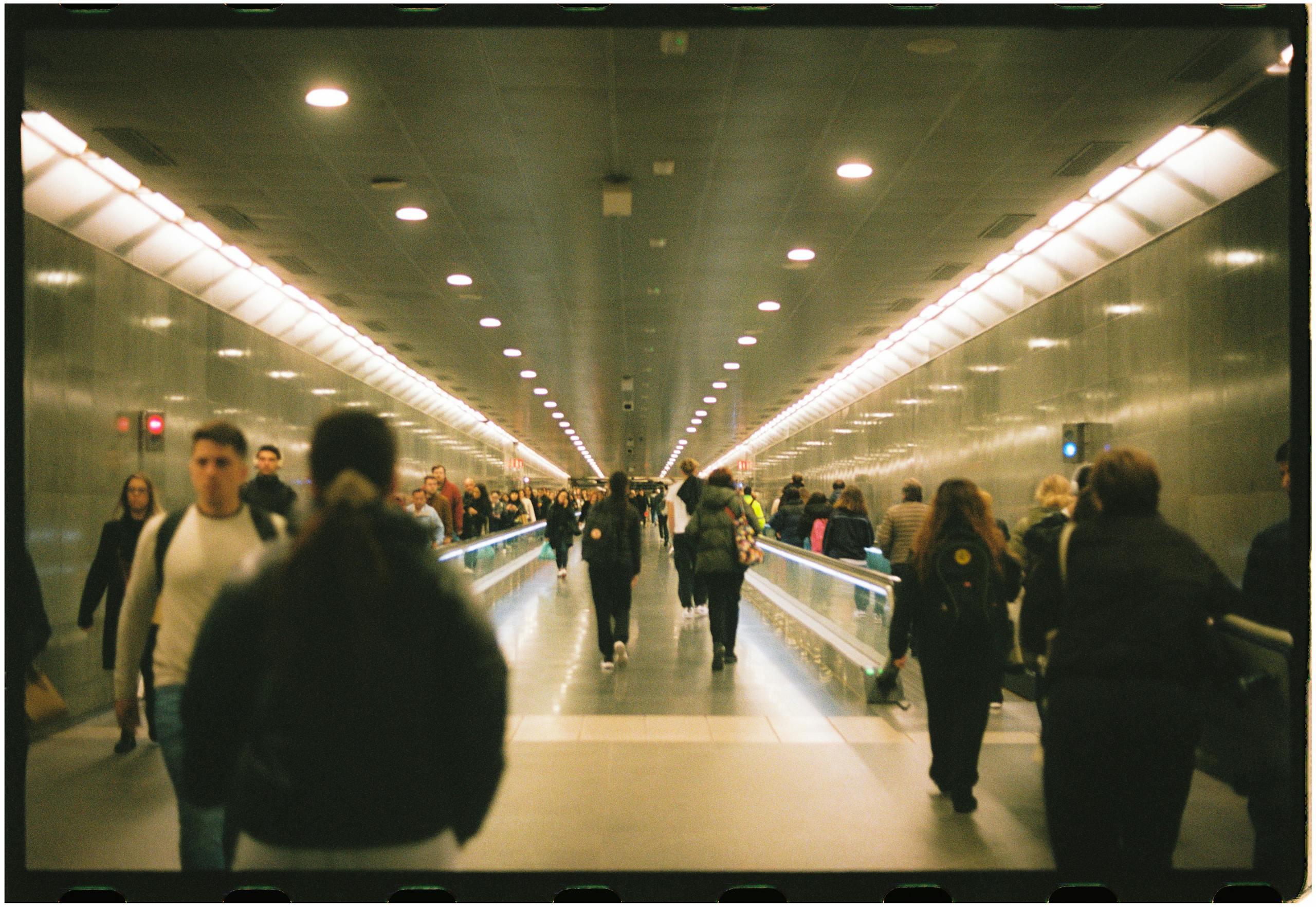 Crowd of travelers walking through an airport terminal with bright lights and a long corridor.